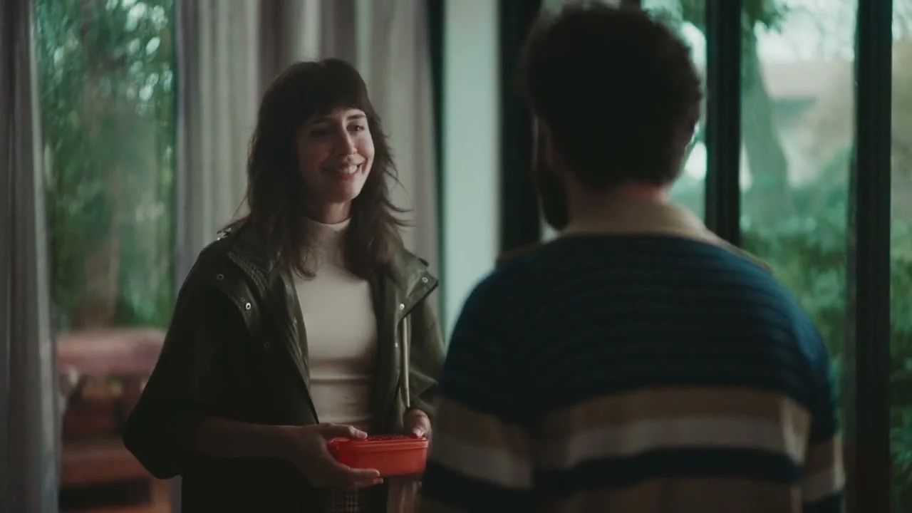 A woman smiling while holding a red container, engaging with a man in a cozy indoor setting with large windows and greenery in the background.