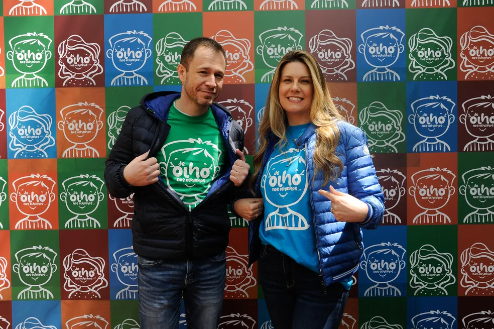 Two smiling individuals pose in front of a colorful backdrop featuring cartoonish faces and the phrase "De Olho Nos Olhinhos." The man wears a green t-shirt and a black jacket, while the woman wears a blue t-shirt and a blue jacket, both showcasing their support for the event.