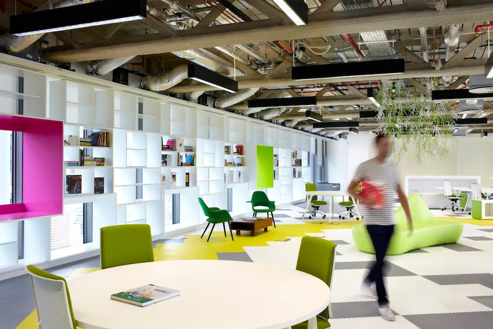 Modern office interior featuring colorful furniture, including green chairs and a pink bookshelf, with a spacious layout and natural lighting. A person is seen walking through the workspace, which includes areas for collaboration and individual work.