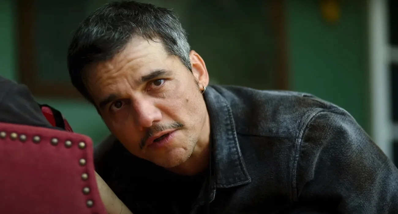 Man with questioning expression wearing a denim jacket leaning on a red chair.