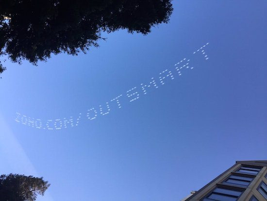 Skywriting message reading "SDCCOM OUTSMART" against blue sky with tree and building corner in view.