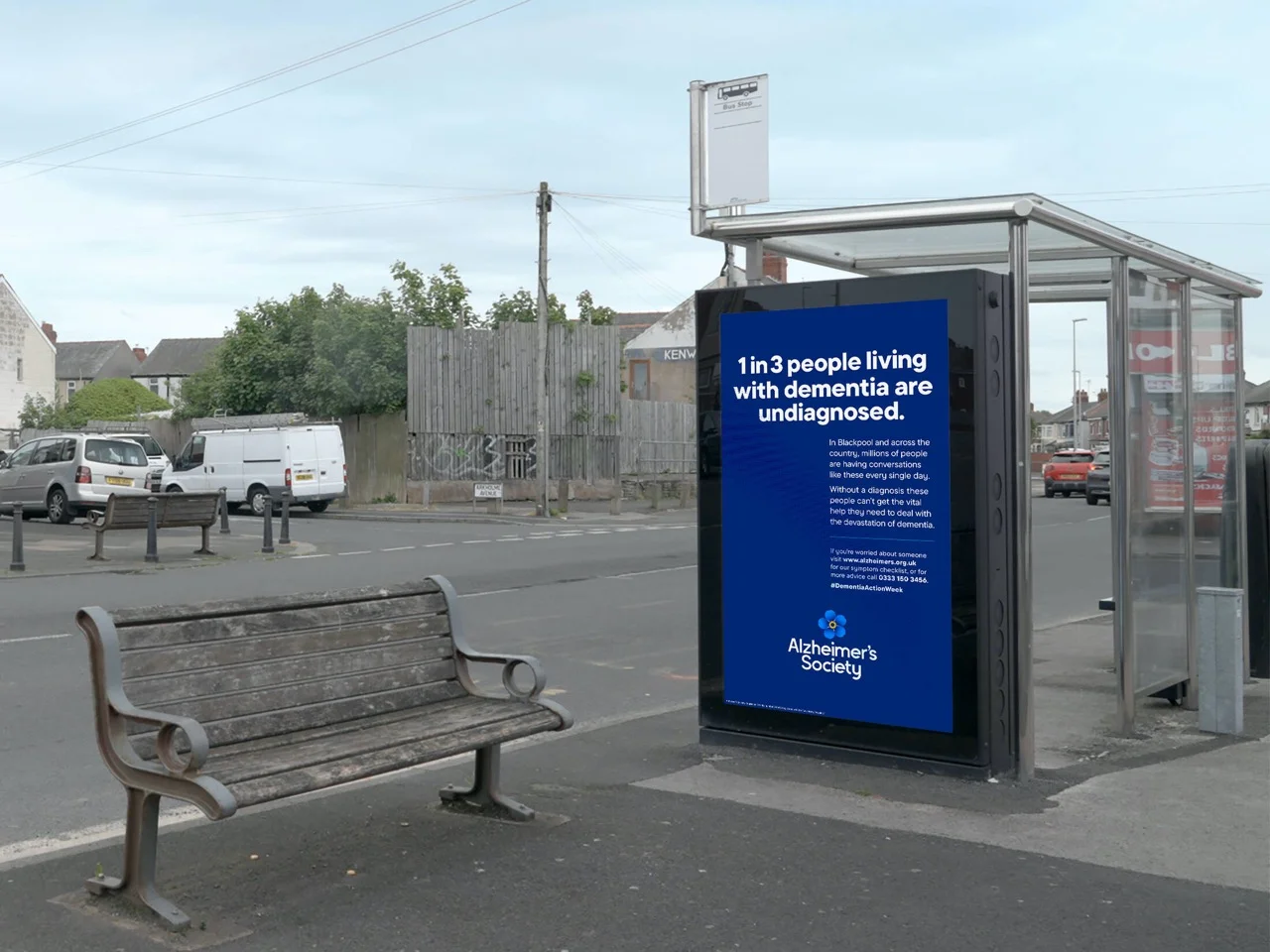 Bus stop with Alzheimer's Society awareness poster about dementia diagnosis next to a wooden bench on a quiet street.