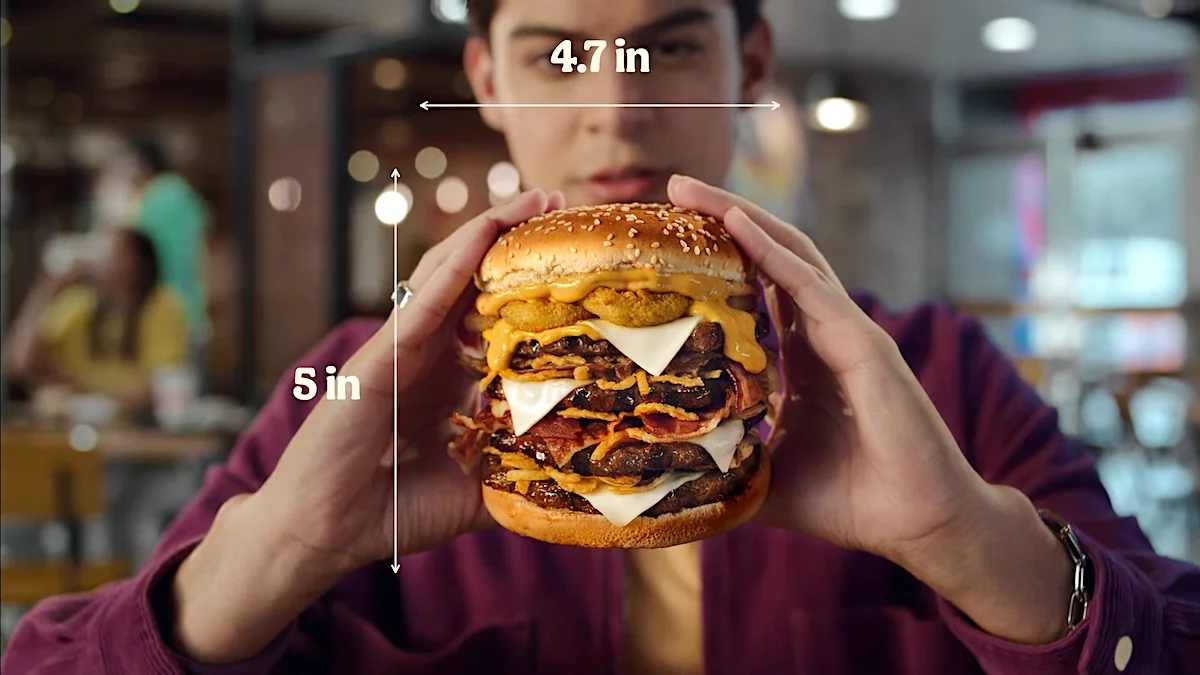 Person holding a large cheeseburger with measurements of 4.7 inches wide and 5 inches tall, featuring multiple layers of beef, cheese, bacon, and sauce in a restaurant setting.