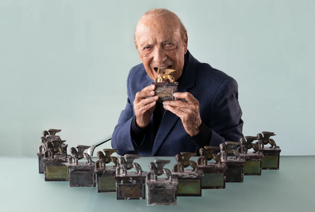 Elderly man in a suit holding and playfully biting a golden trophy, surrounded by multiple identical trophies on a table.