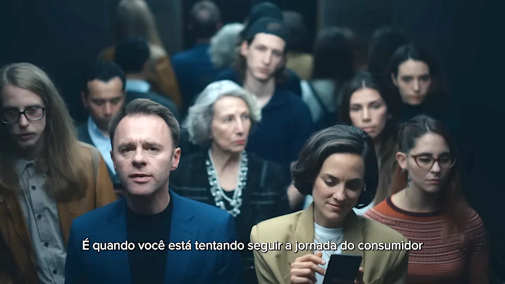 Group of people in an elevator, with a man speaking and a woman using a smartphone, Portuguese subtitle about consumer journey. FOMA
