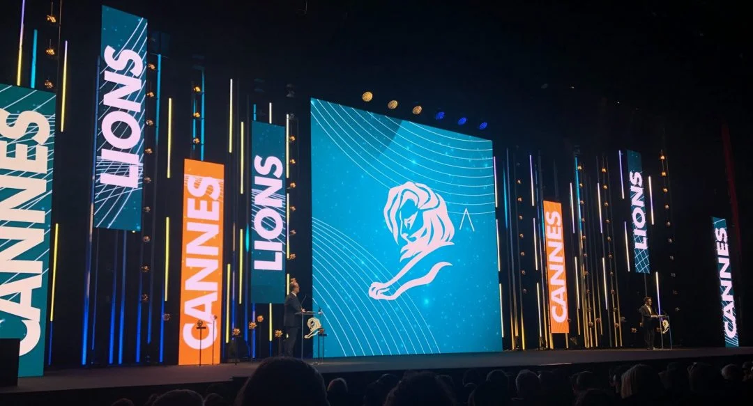 Cannes Lions stage with large screens displaying the event name and logo, featuring a speaker at the podium.