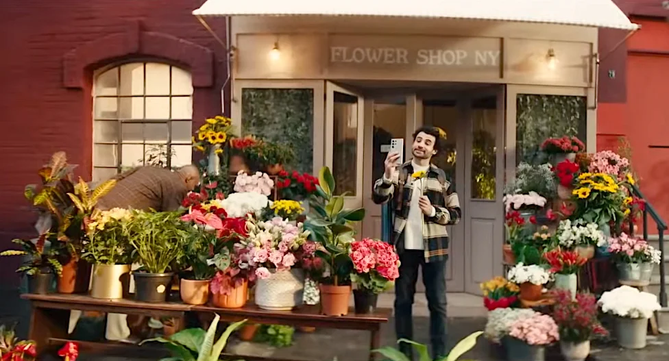 Man taking a selfie in front of a flower shop in New York with a variety of colorful flowers displayed outside.