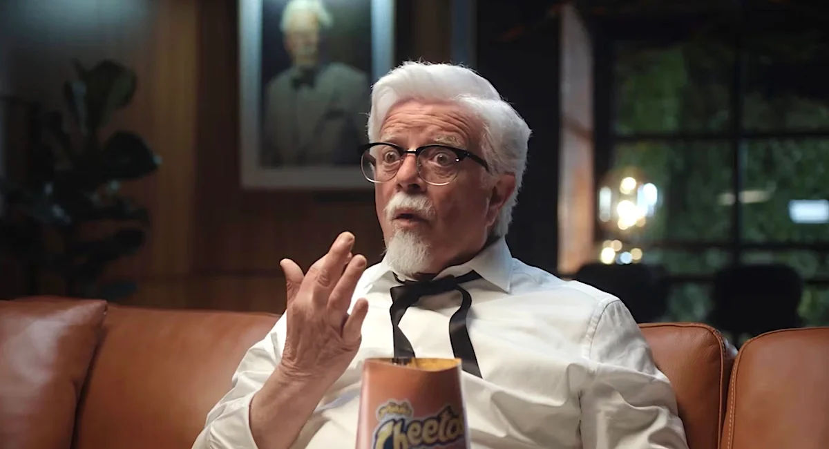 Elderly man with white hair and glasses eating Cheetos on a leather couch in a dimly lit room.