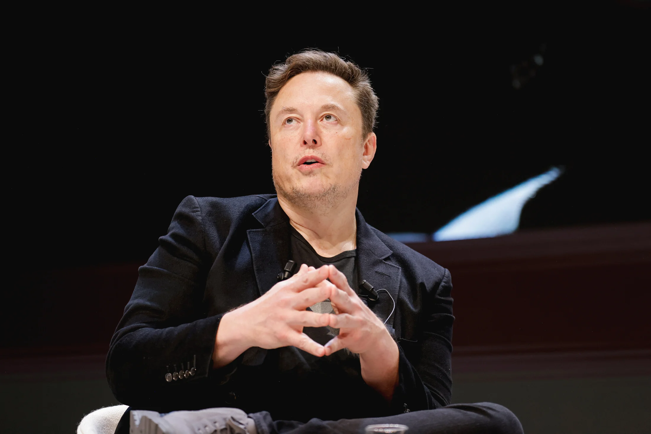 Man in black suit speaking at a conference, hands clasped, dark background.