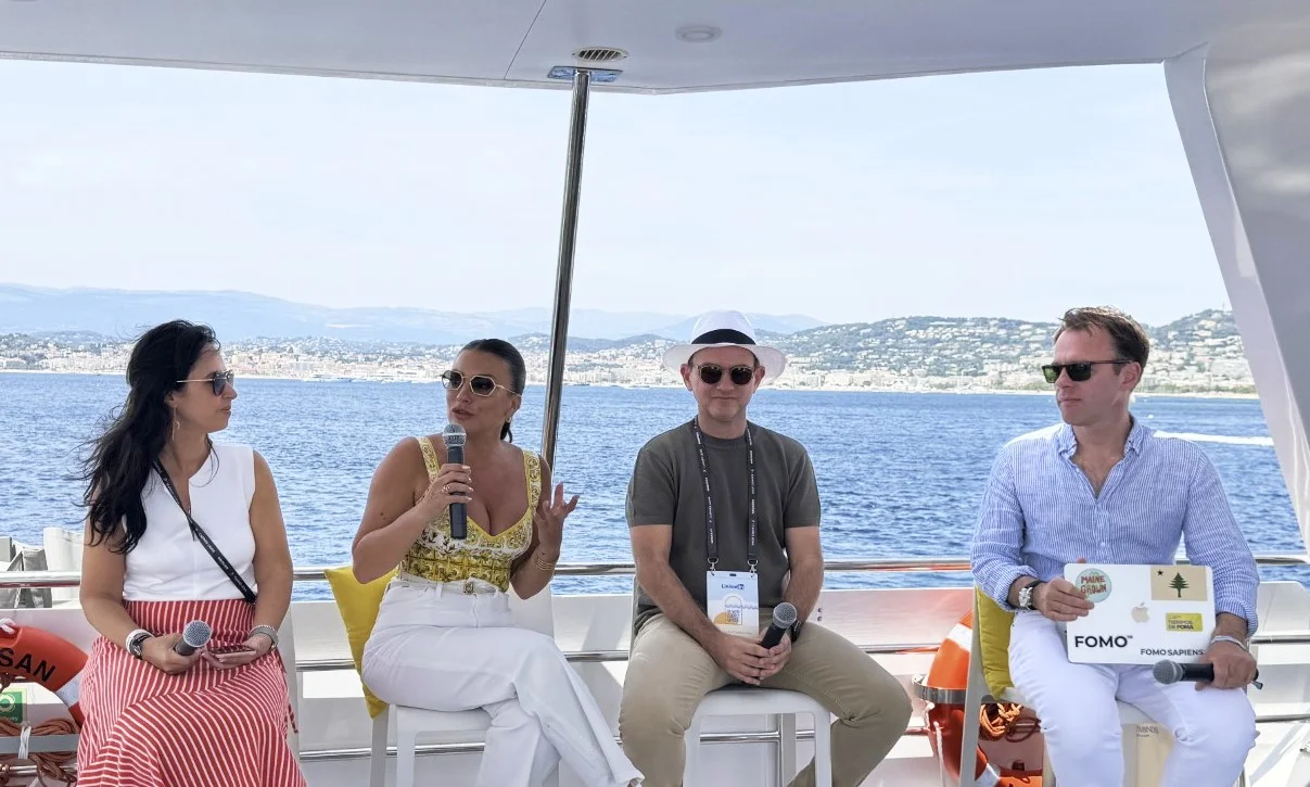 8 Tendências de Marketing. Panel discussion on a boat with four people holding microphones, ocean and coastal town in the background.