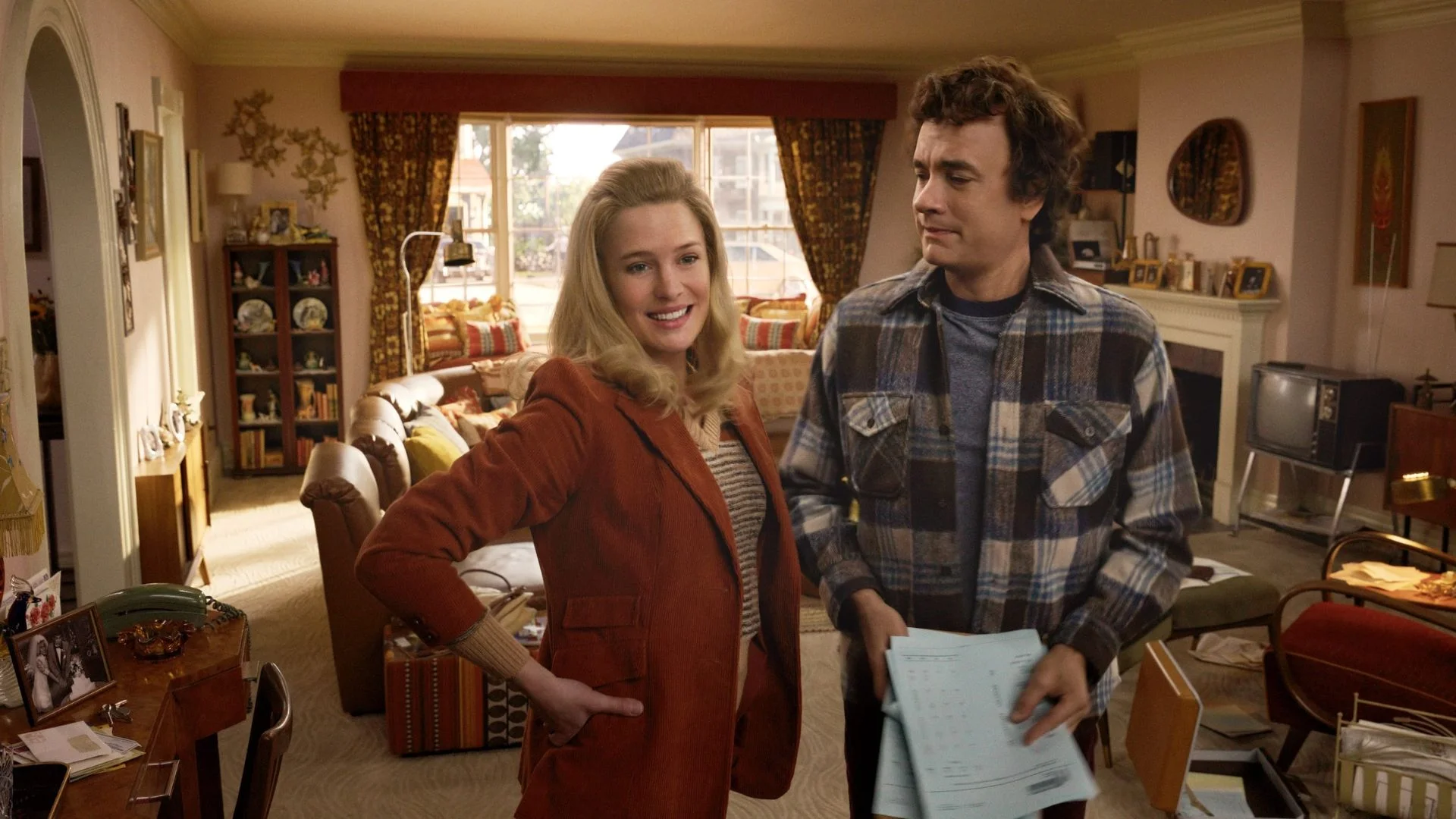 Two people standing in a cozy living room, one smiling and the other holding papers, vintage decor in the background.