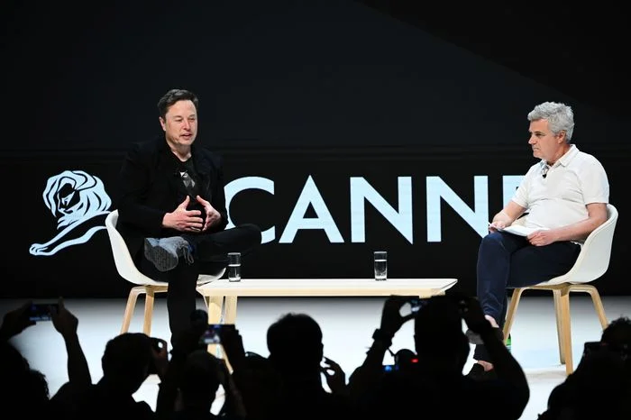 Two men seated on stage during a discussion at Cannes Lions International Festival of Creativity, with audience members taking photos.