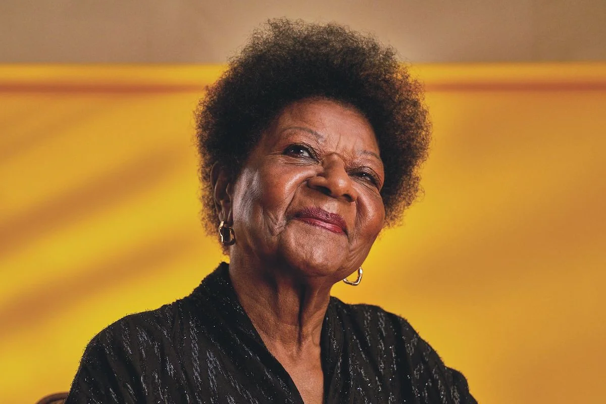 Elderly woman with curly hair and earrings smiling against a yellow background.