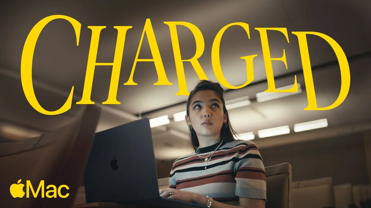 Woman using a MacBook in a dimly lit room with the word "CHARGED" in large yellow text above her