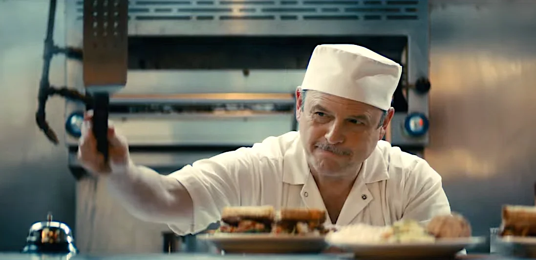 Chef in a white uniform and hat holding a spatula in a professional kitchen with sandwiches and plates of food in the foreground