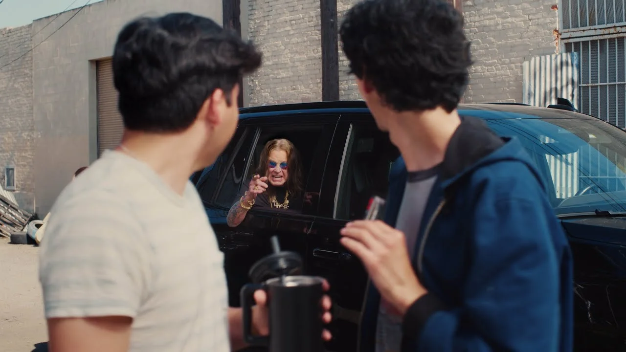 Two young men looking at an older man with long hair and sunglasses leaning out of a car window in an urban setting.