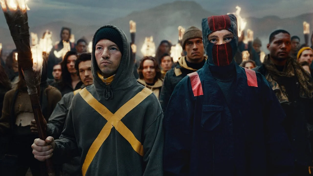 Group of people holding torches, wearing hooded and masked outfits, standing in a dark, mountainous setting.