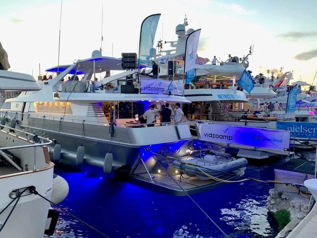 Luxury yachts docked at a marina during sunset with people socializing on decks, illuminated with blue lights.