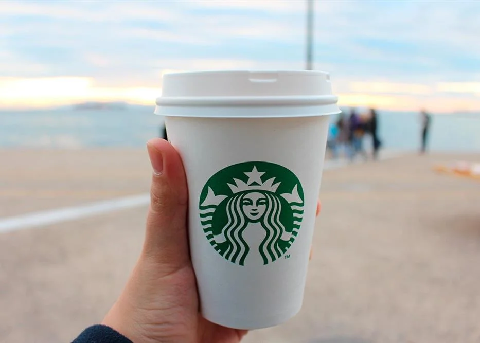 Logo Starbucks. Hand holding a Starbucks coffee cup with a beach background.