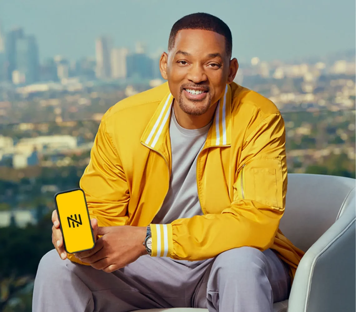 Will Smith falando em português. Man in yellow jacket holding smartphone with yellow screen displaying logo, cityscape background.