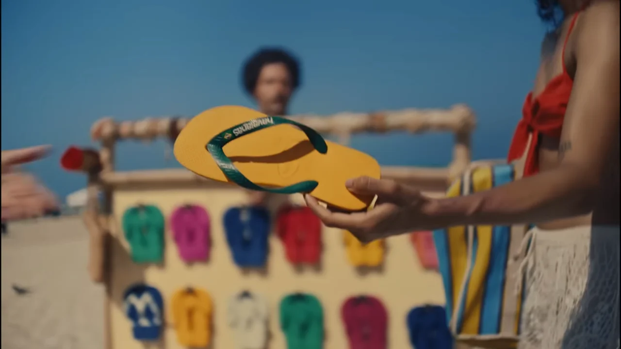 Revezamento de Havaianas. A hand holding a vibrant yellow flip-flop with a green strap, set against a sunny beach backdrop featuring a display of colorful flip-flops in various shades.