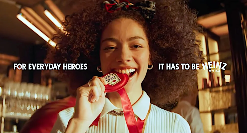 Woman smiling and biting a Heinz ketchup medal with the text "For Everyday Heroes" and "It Has to Be Heinz" in the background.