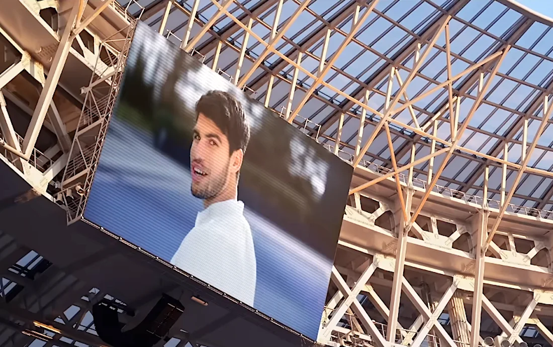 Large digital screen displaying a man in a white shirt inside a modern stadium with a glass roof and metal framework.