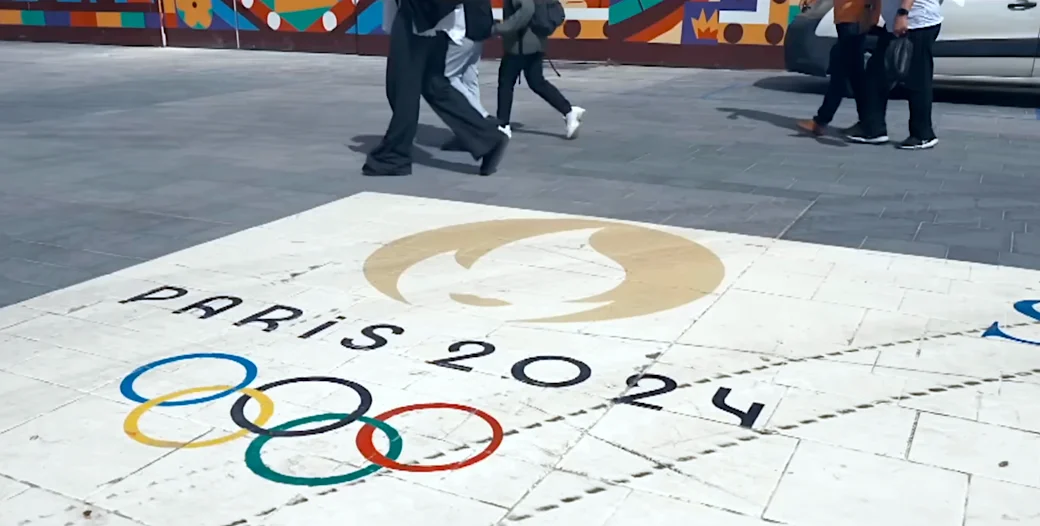 Pedestrians walking over the Paris 2024 Olympic Games logo and rings on a tiled pavement, showcasing the vibrant atmosphere of the upcoming event.