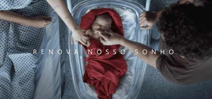 Newborn baby wrapped in red blanket in hospital crib with parents' hands touching, text Renova Nosso Sonho.