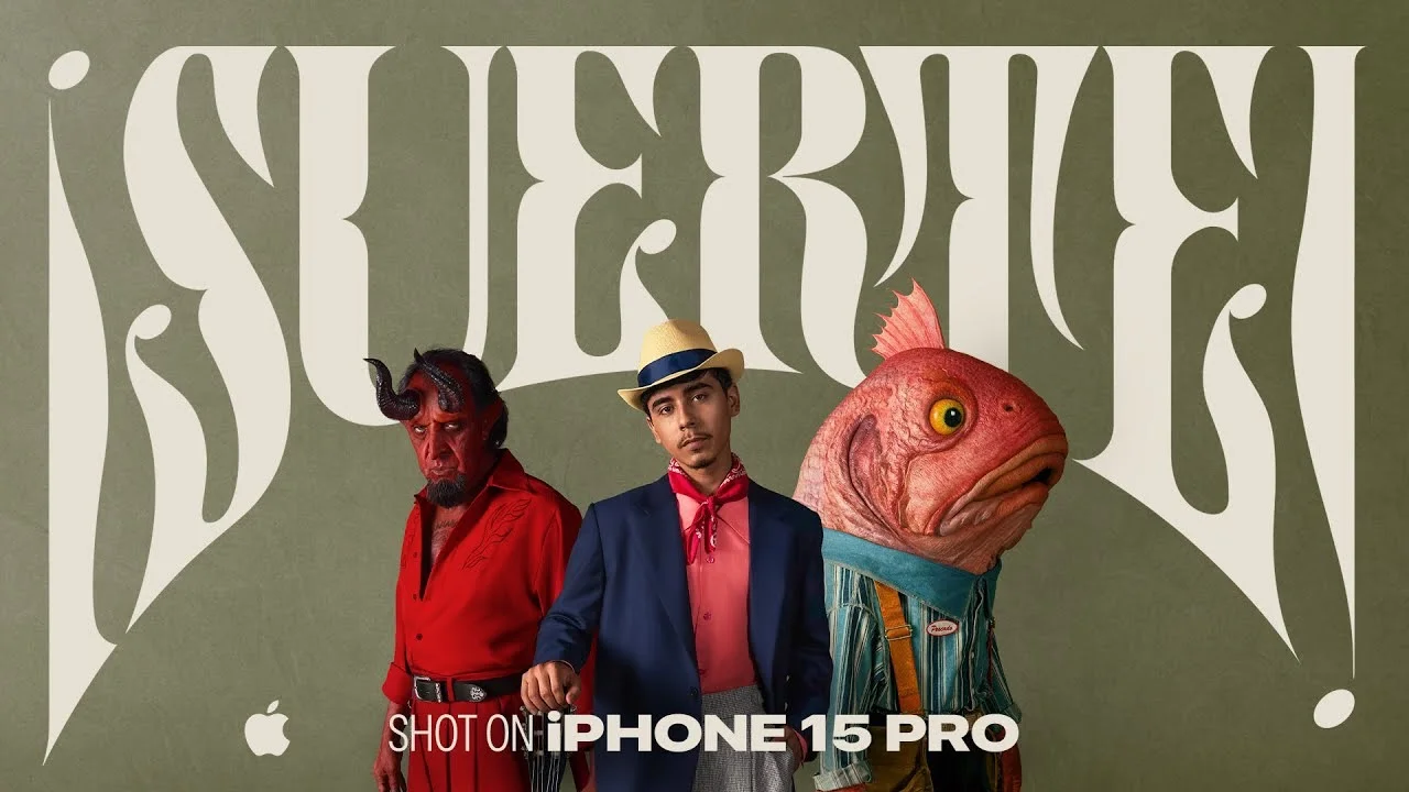 Three characters in colorful costumes, including a devil, a man in a hat, and a fish-headed person, standing in front of stylized text "Suerte," promoting the iPhone 15 Pro.