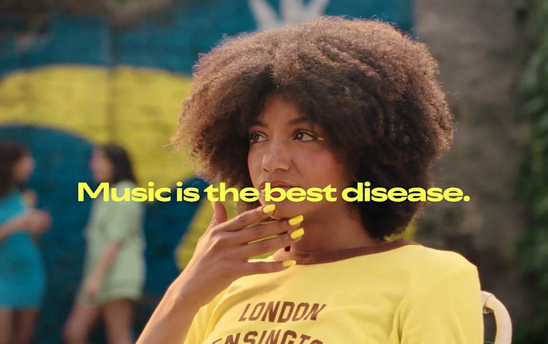 Woman with curly hair and yellow nails wearing a yellow shirt that says London Kensington, with the text Music is the best disease in the foreground.