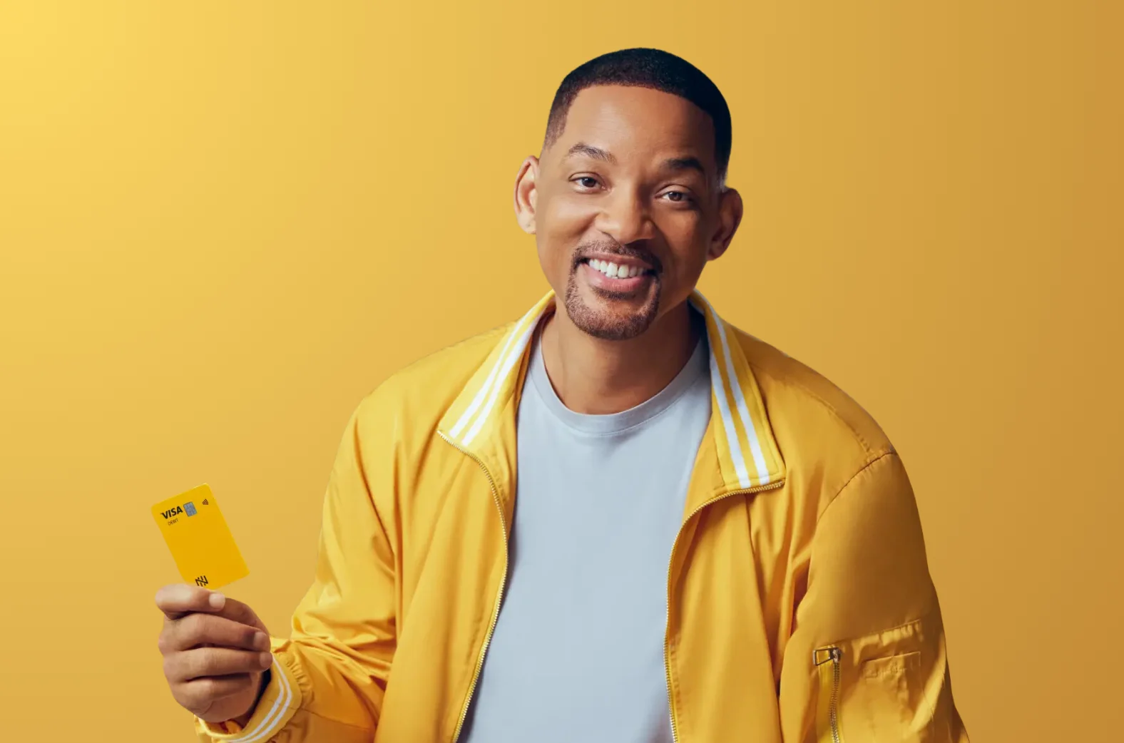 Smiling man in a yellow jacket holding a yellow Visa card against a yellow background.