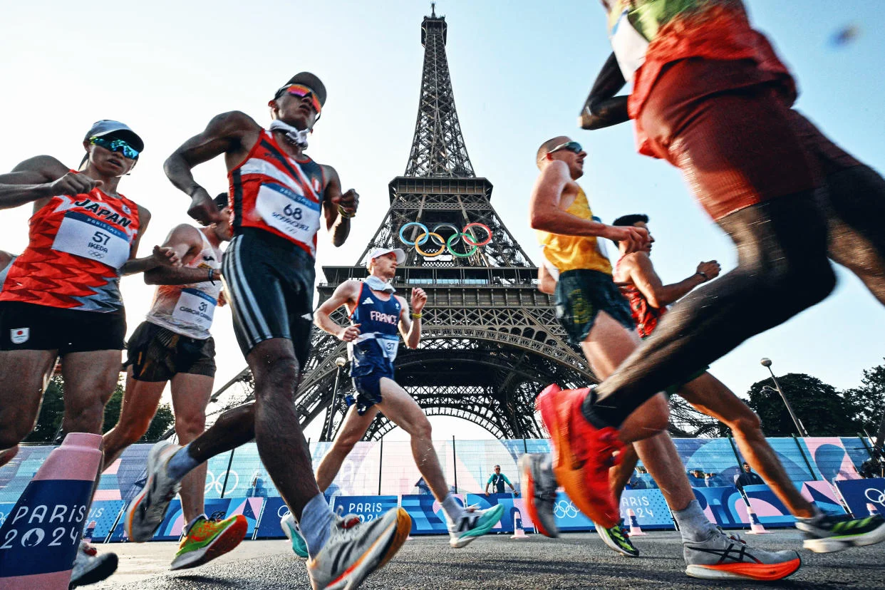 Athletes competing in a race near the Eiffel Tower during the Paris 2024 Olympics, showcasing diverse national uniforms and the iconic Olympic rings in the background.