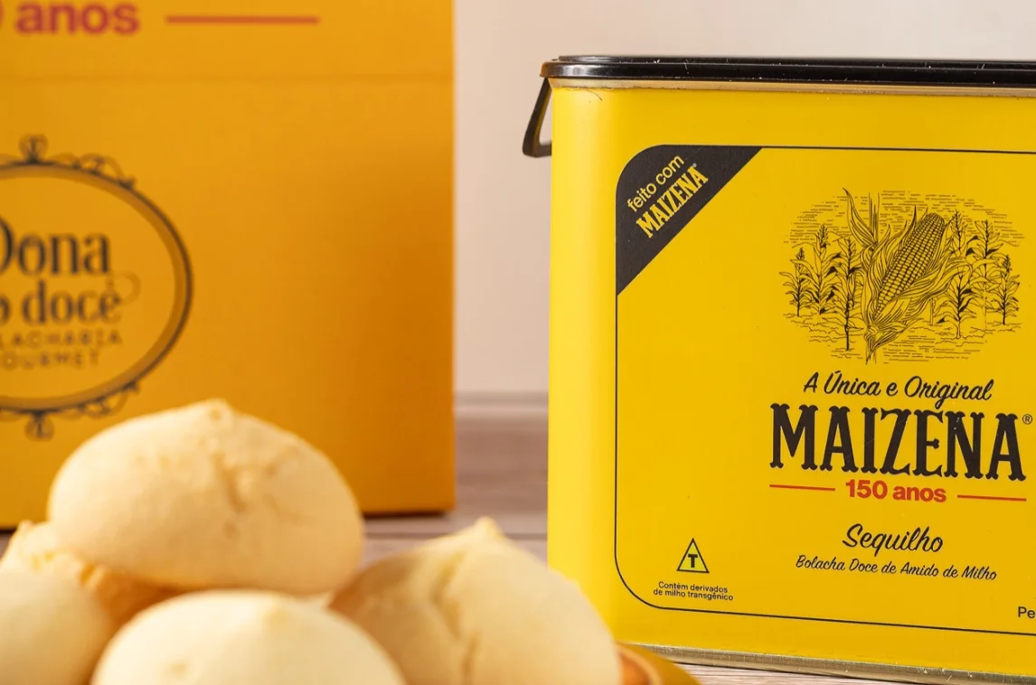 Maizena e Dona do Doce. Yellow Maizena flour canister with a vintage design, featuring the text "A Única e Original MAIZENA 150 anos," next to a box labeled "Dona Doce." In the foreground, there are sweet cornmeal cookies.