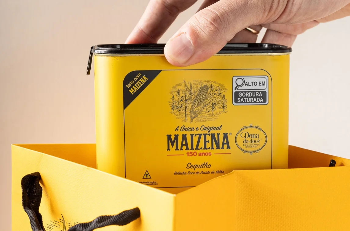 Hand reaching for a yellow tin of Maizena, a corn starch product, inside a matching yellow shopping bag, highlighting its 150-year heritage and gourmet baking use.