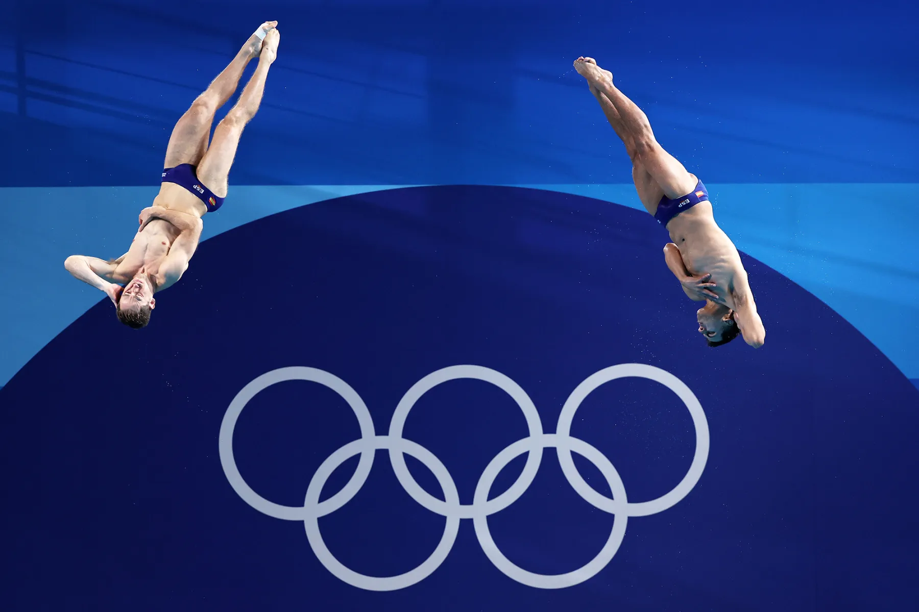 Two male divers performing synchronized dives during the Olympics, with the Olympic rings logo prominently displayed in the background.