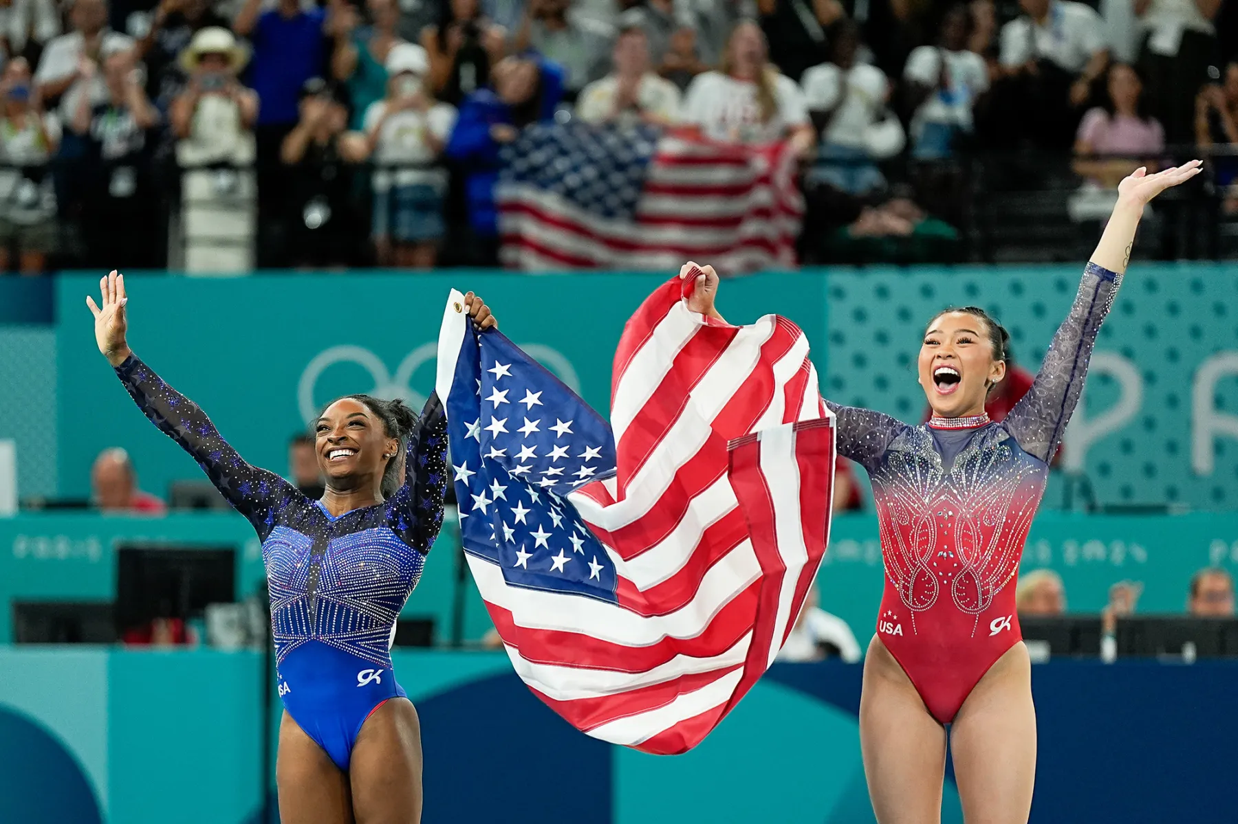 Two female gymnasts celebrate at the Olympics, joyfully holding a large American flag. They are wearing colorful, sparkly leotards and are surrounded by a cheering crowd, showcasing a moment of national pride and athletic achievement.