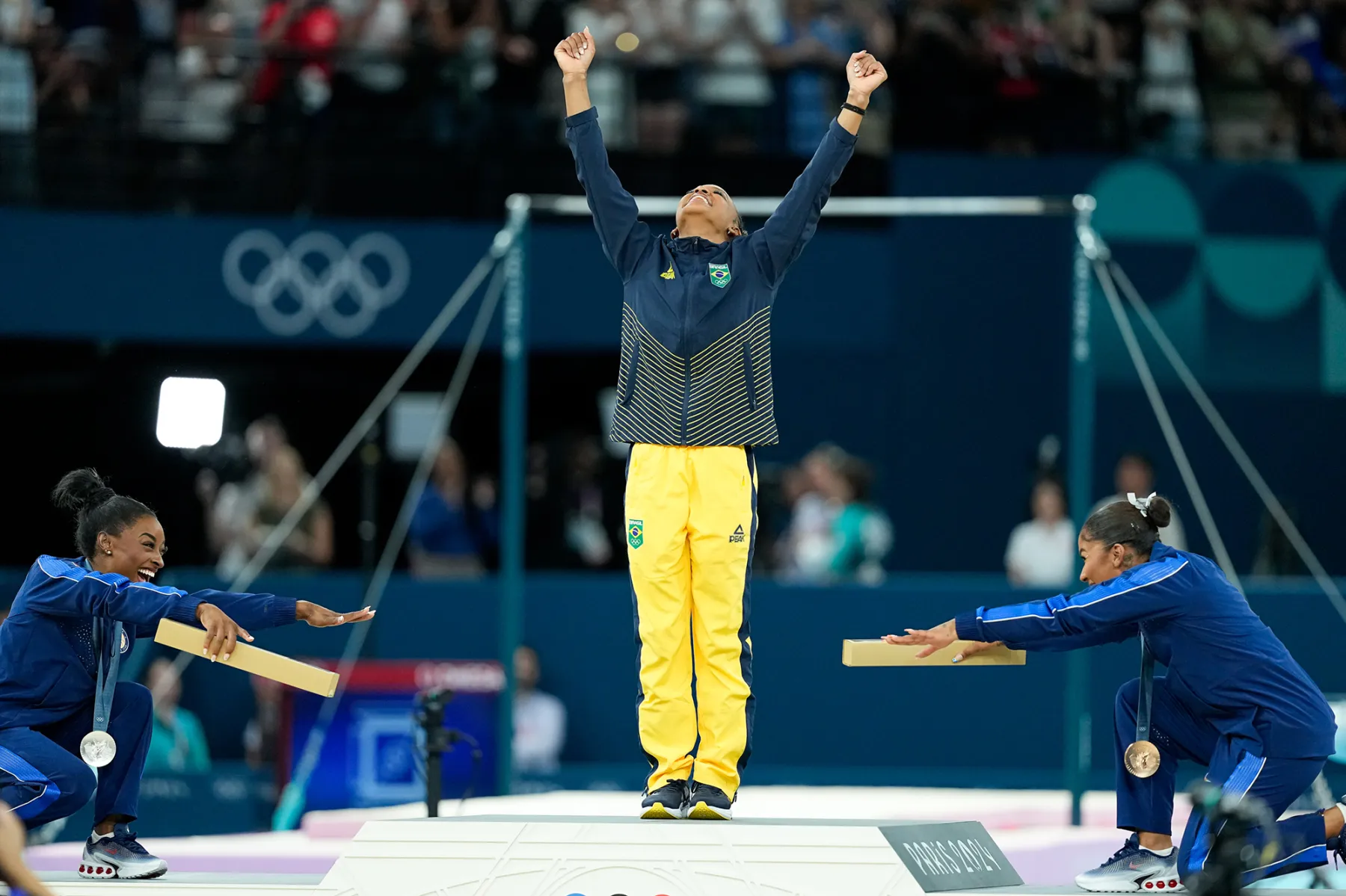 A Brazilian gymnast celebrates on the podium at the Olympics, raising arms in triumph, while teammates with medals cheer and support her from the side. The event showcases the spirit of sportsmanship and achievement in gymnastics.