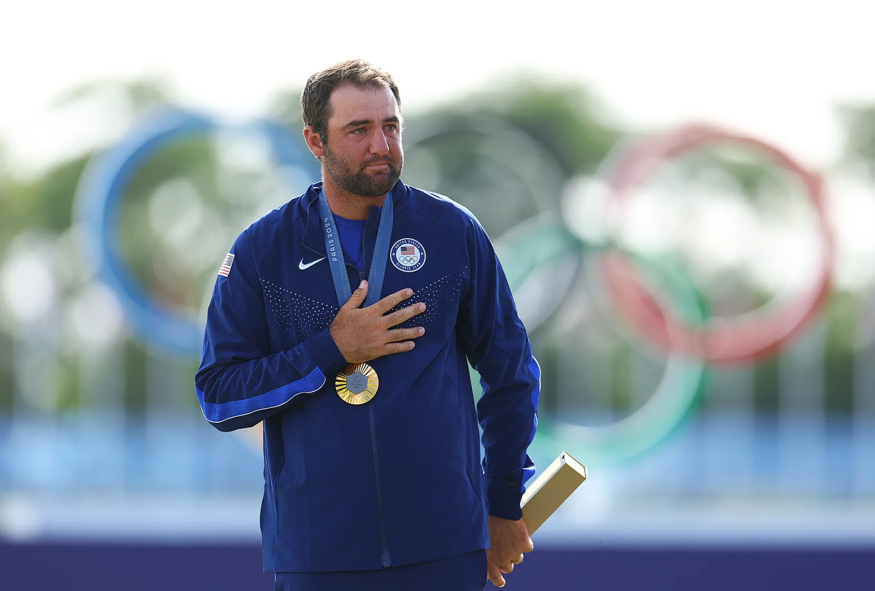 Athlete wearing a blue jacket holds a gold medal and a trophy, standing in front of the Olympic rings, showcasing pride and achievement at the Olympic Games.