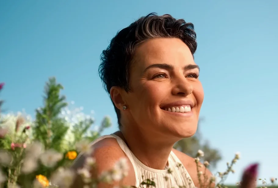 Smiling woman with short dark hair enjoying a sunny day surrounded by colorful wildflowers against a clear blue sky.