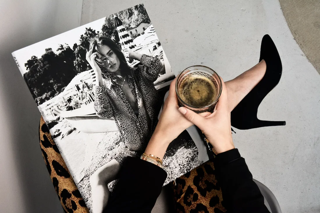 Woman holding a glass of drink while seated, wearing stylish black high heels and a leopard print outfit, with a fashion magazine featuring a black-and-white photo of a model placed on her lap.