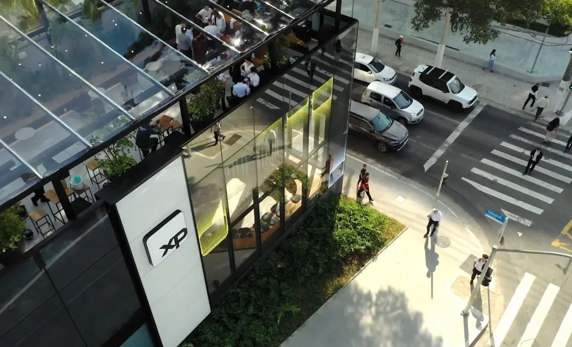 Aerial view of the modern XP office building featuring a glass facade, outdoor seating area, and people engaging in discussions, set against a busy city street with cars and pedestrians.