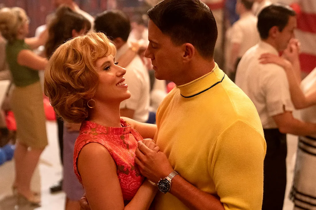 Couple dancing closely in a vibrant retro-themed setting, featuring a woman in a floral dress and a man in a bright yellow sweater, capturing a romantic moment amidst a lively dance floor.