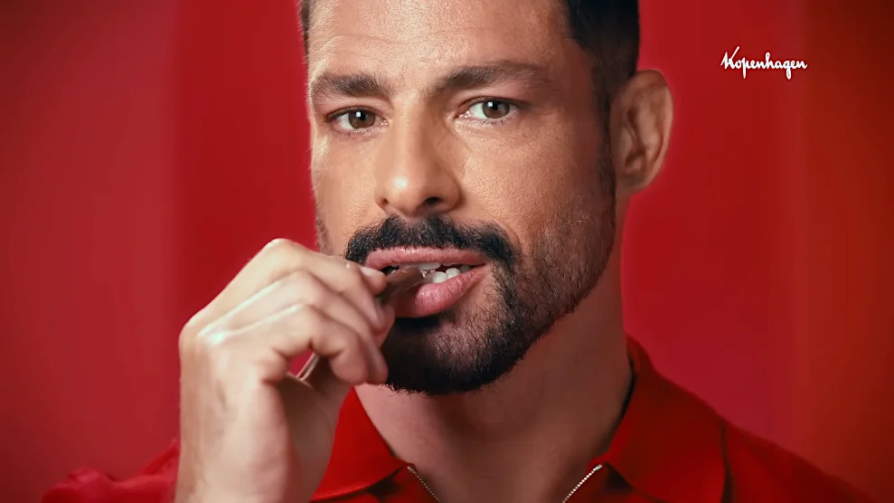 Close-up of a man with a beard and striking green eyes, wearing a red shirt, holding a small object near his lips against a vibrant red background, promoting Copenhagen products.