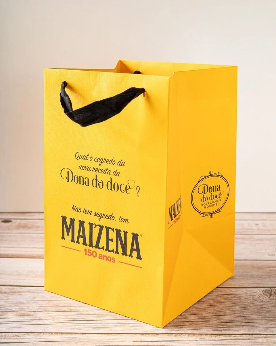 Yellow shopping bag featuring "Dona do doce" branding and "MAIZENA 150 anos" text, placed on a wooden surface.