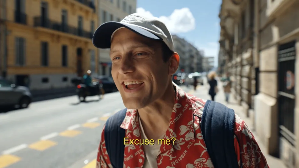 A man in a floral shirt and cap smiles while speaking on a busy street, with pedestrians and vehicles in the background, capturing a lively urban atmosphere.