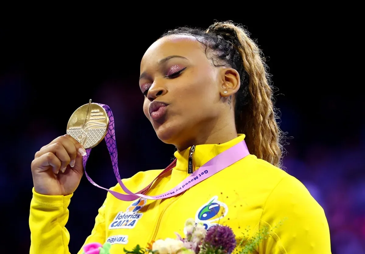 Athlete celebrating with a gold medal, wearing a yellow tracksuit and pink ribbon, showcasing a joyful expression and holding a bouquet of flowers.