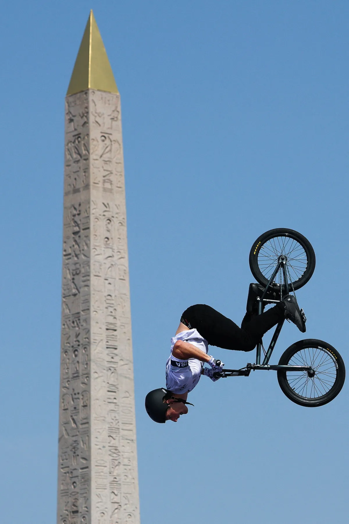 BMX rider performing a backflip stunt against a clear blue sky, with an ancient Egyptian obelisk in the background, showcasing extreme sports and cultural landmarks.