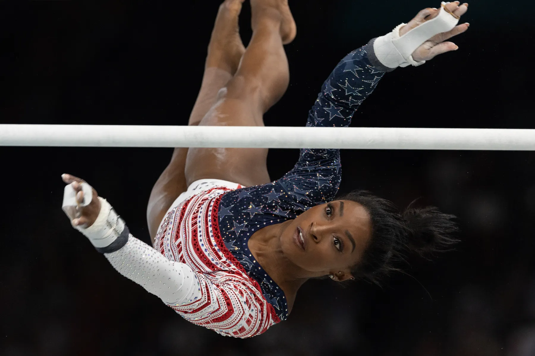 Gymnast performing a dynamic move on the uneven bars, showcasing athleticism and grace in a patriotic-themed leotard adorned with stars and stripes.