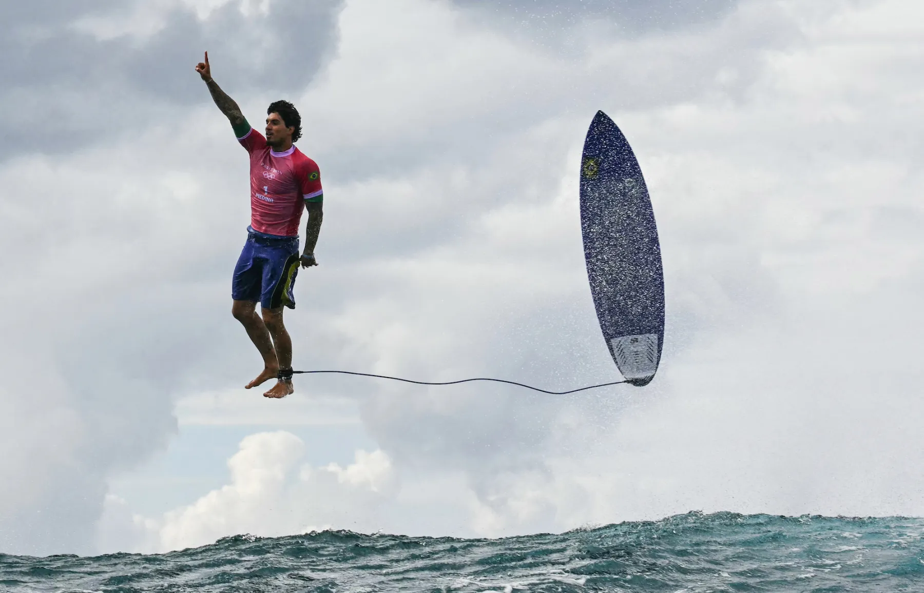 melhores fotos das Olimpíadas 2024. Surfer performing an aerial maneuver above the waves while holding onto a surfboard, with a dramatic sky and ocean backdrop.